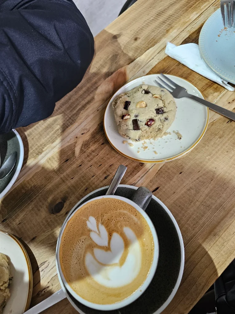 Taza de cerámica con tulip latte art perfecto y galleta artesanal de avena y chocolate en mesa de madera nativa.